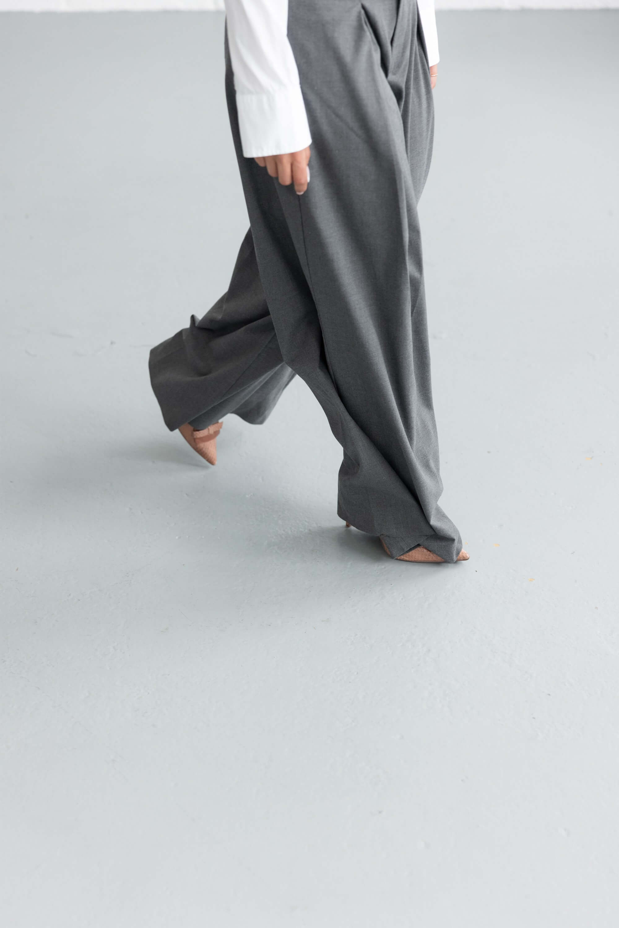 A woman wearing grey tailored suit trousers walks in pointed pale pink stiletto heels.