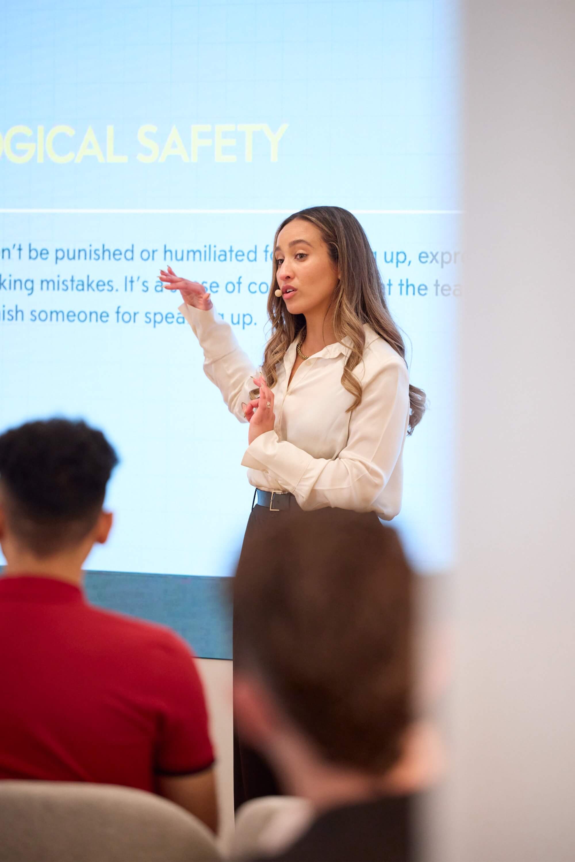Amira presenting to a crowd. She is standing in front of a projector with one arm slightly raised in explanation. She wears a white shirt and talks into a headset microphone.