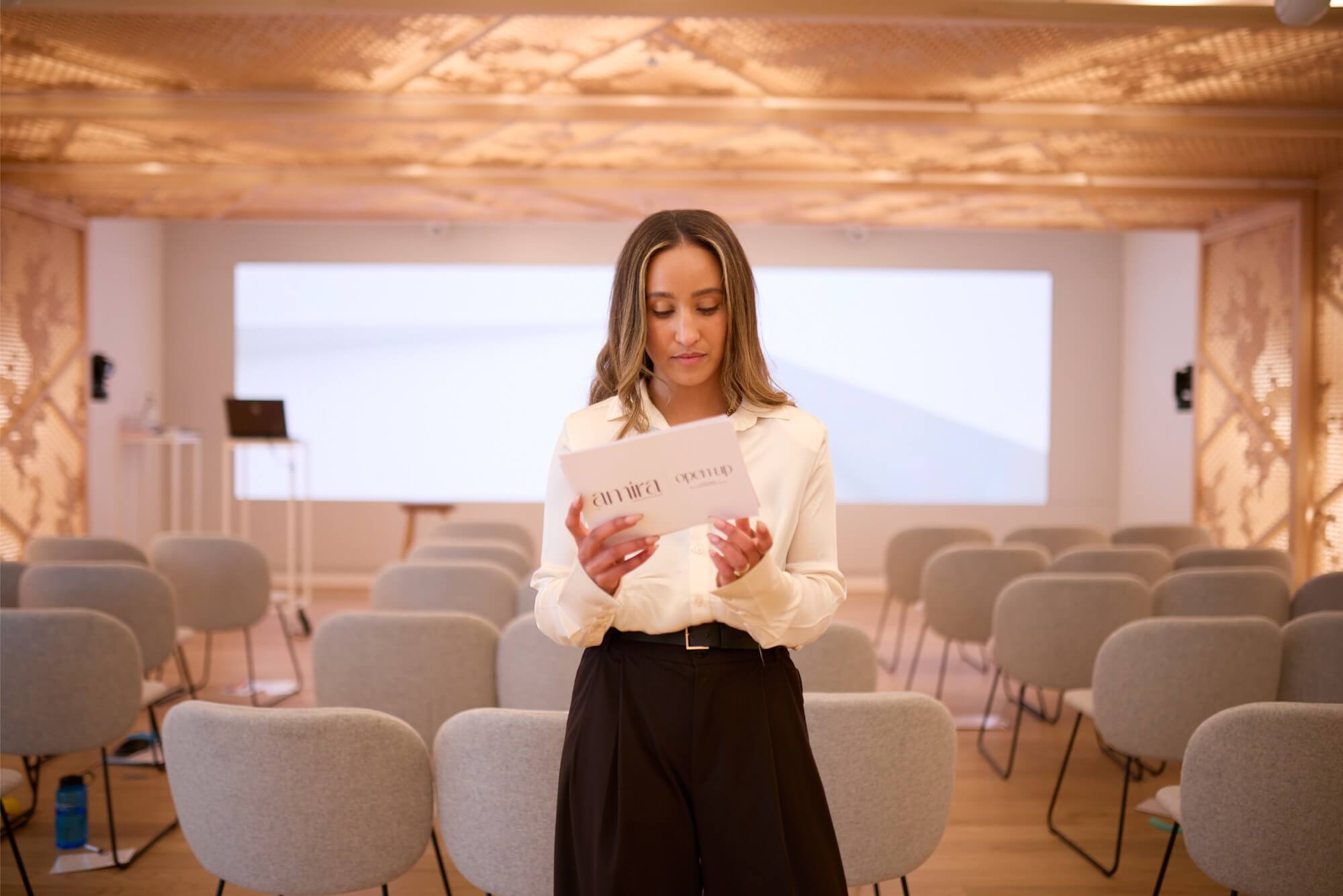 Amira holds and looks down at some presentation note cards. She stands in front of a number of empty chairs and a projector.
