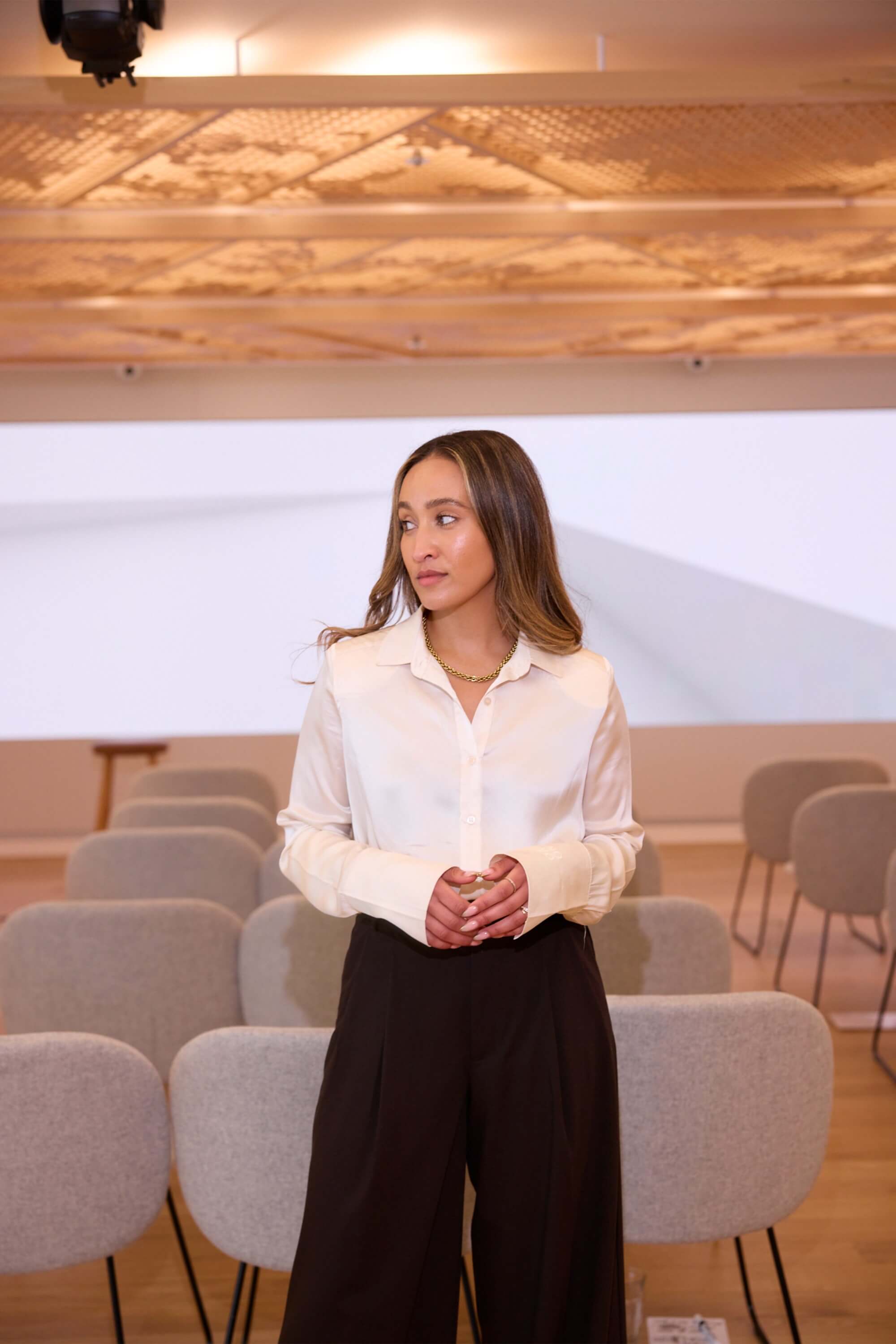 Amira stands in a white shirt and black trousers in front of a row of empty chairs and projector.